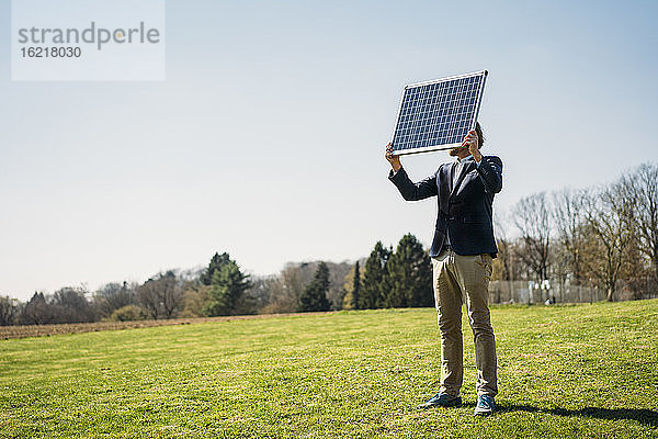 Männlicher Fachmann  der ein Solarmodul hält  während er im Park auf dem Rasen steht  gegen einen klaren Himmel an einem sonnigen Tag
