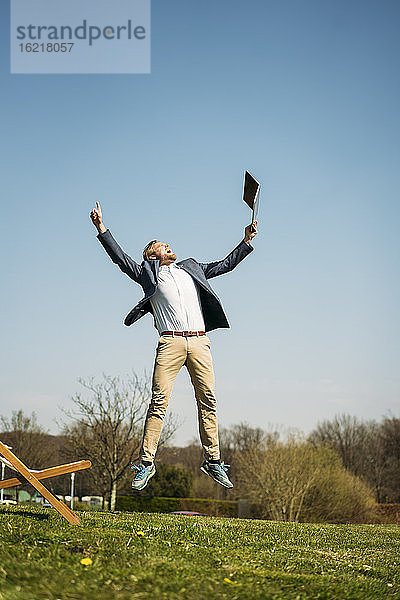 Aufgeregter Geschäftsmann  der einen Laptop hält  während er im Park gegen den klaren Himmel an einem sonnigen Sommertag auf das Gras springt
