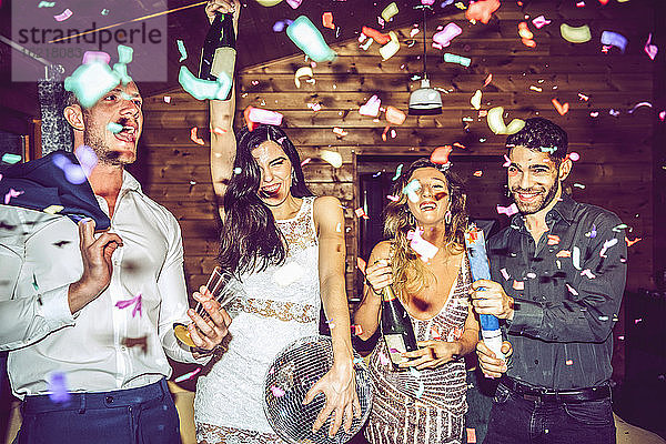 Männliche und weibliche Freunde mit Champagner und Discokugel tanzen inmitten von Konfetti in der Partei