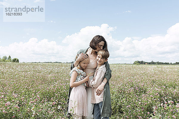 Liebevolle Mutter  die ihre Kinder umarmt  während sie inmitten von Blumen im Feld vor dem Himmel steht