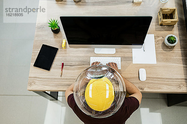 Geschäftsmann trägt ein Fischglas im Kopf  während er einen Computer in einem modernen Büro benutzt