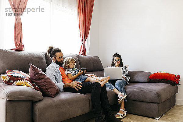 Frau arbeitet am Laptop  während Vater und Tochter auf dem Sofa zu Hause ein Smartphone benutzen