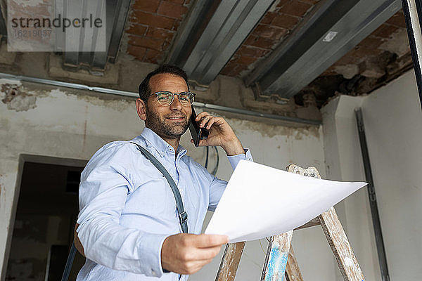 Porträt eines selbstbewussten Architekten mit Plan am Telefon in einem im Bau befindlichen Haus