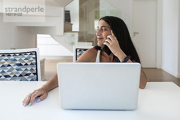 Lächelnde Frau mit Laptop auf dem Tisch  die über ihr Mobiltelefon spricht  während sie zu Hause sitzt