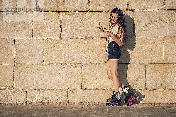 Junge Frau fährt Inline-Skates und lehnt sich mit ihrem Smartphone an eine Wand