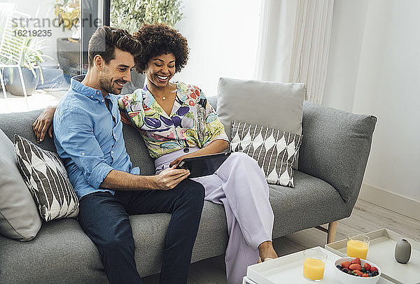 Ein glückliches Paar benutzt ein digitales Tablet  während es auf dem Sofa im Wohnzimmer eines Penthouses sitzt.