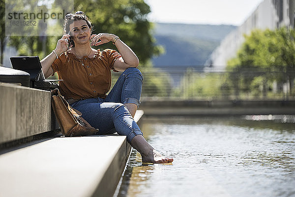 Lächelnde Frau  die über ihr Handy spricht  während sie an einem sonnigen Tag am Teich im Park sitzt