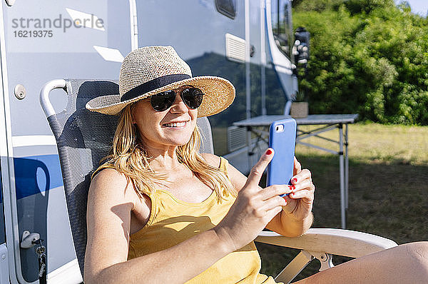 Entspannte Frau  die neben einem Wohnmobil sitzt  ihren Urlaub genießt und ihr Smartphone benutzt