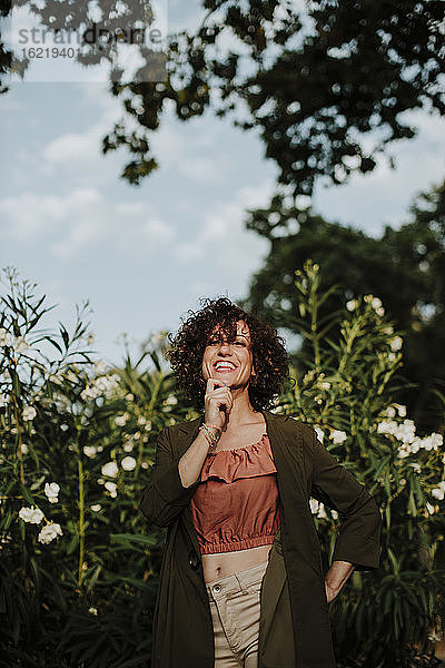 Lächelnde mittelgroße erwachsene Frau mit lockigem Haar und Mantel  die gegen Pflanzen im Park steht