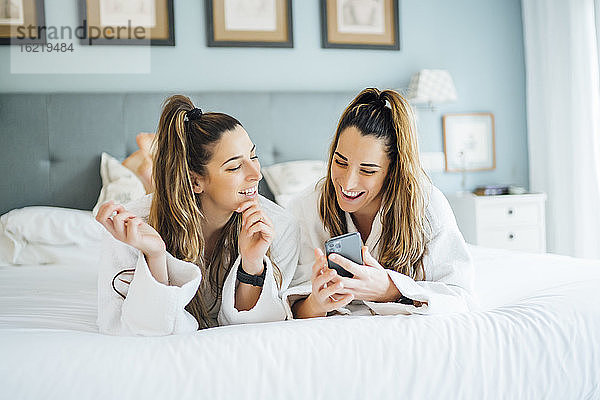 Lächelnde Zwillingsschwestern  die ein Smartphone benutzen  während sie auf dem Bett im Hotel liegen