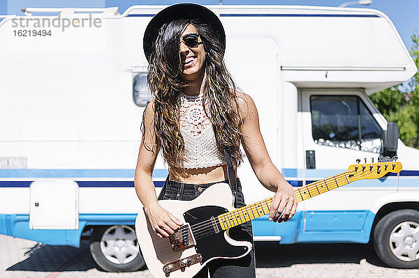 Glückliche junge Frau spielt E-Gitarre vor einem Wohnmobil