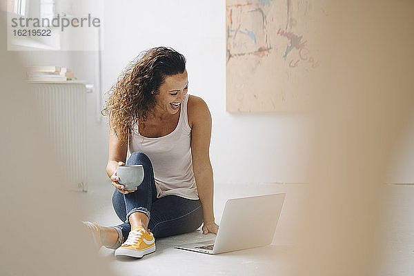 Fröhliche Frau  die einen Kaffeebecher hält  während sie einen Laptop auf dem Boden zu Hause benutzt