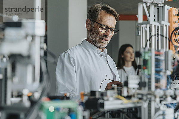 Älterer Mann untersucht Maschinen mit einer Kollegin im Hintergrund in einem Labor