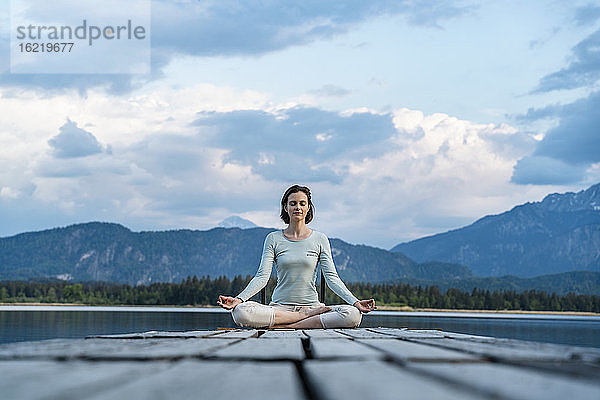 Frau mit geschlossenen Augen meditiert auf Steg über See gegen bewölkten Himmel