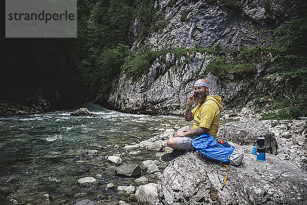 Wanderer mit Vollbart und gelbem Kapuzenpulli sitzt auf einem Stein neben dem Fluss und benutzt sein Smartphone