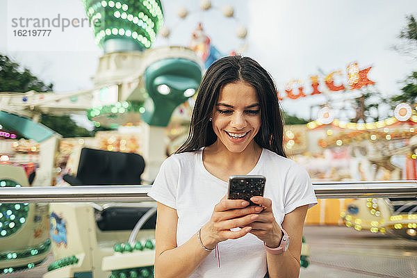 Lächelnde junge Frau  die ein Smartphone benutzt  während sie in einem Vergnügungspark steht
