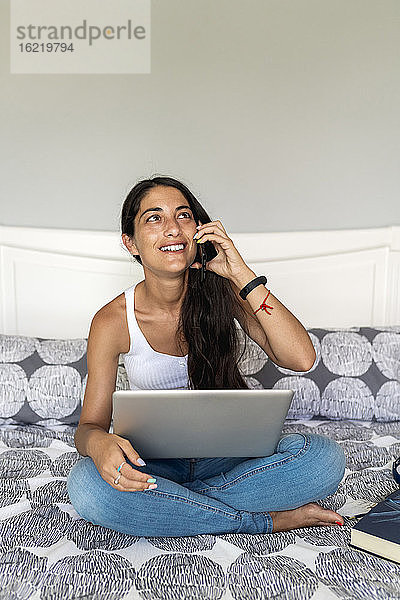 Lächelnde Frau im mittleren Erwachsenenalter  die über ein Smartphone spricht  während sie mit einem Laptop auf dem Bett sitzt