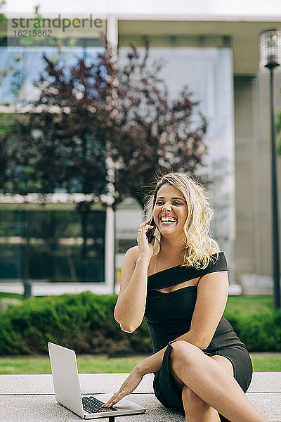 Fröhliche Geschäftsfrau  die mit dem Handy telefoniert und einen Laptop im Freien benutzt