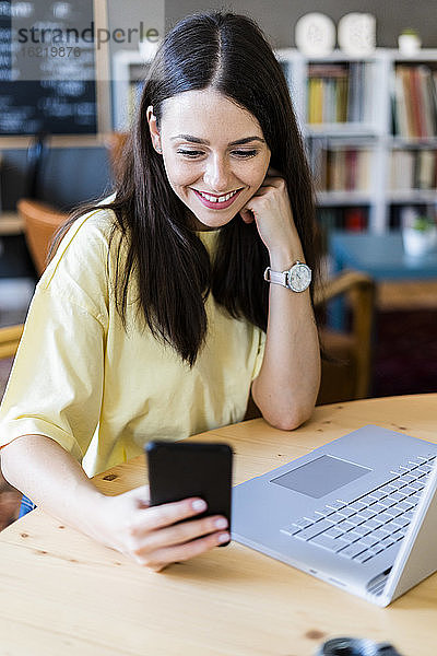 Glückliche Frau  die ein Smartphone benutzt  während sie mit einem Laptop in einem Café sitzt