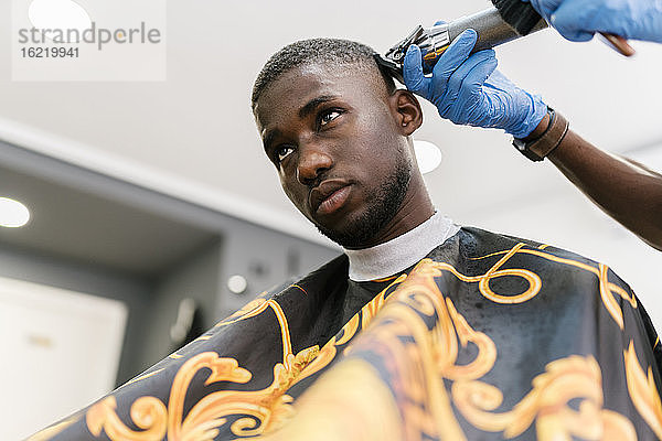 Männlicher Friseur mit Handschuh und Rasiermesser  der einem jungen Mann im Salon die Haare schneidet