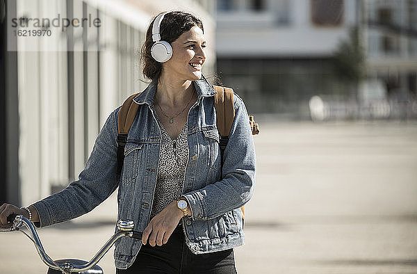 Lächelnde Frau hört Musik über Kopfhörer  während sie mit dem Fahrrad in der Stadt spazieren geht