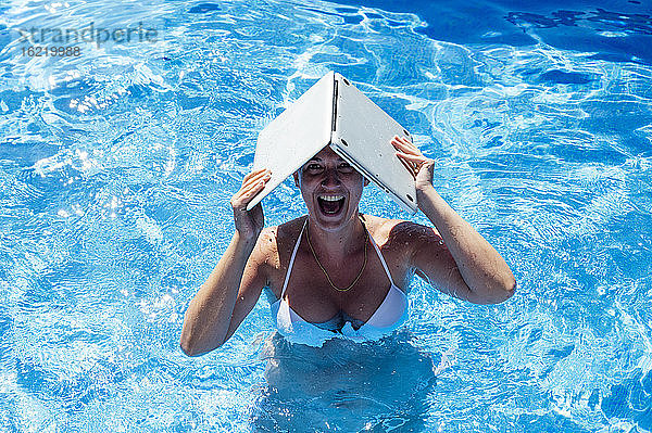 Fröhliche Frau mit Laptop auf dem Kopf beim Schwimmen im Pool