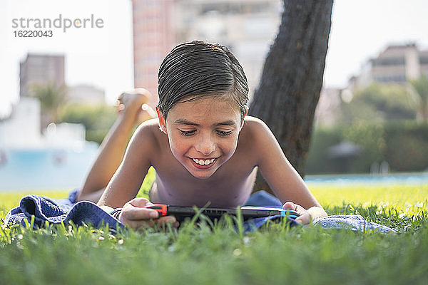Lächelnder Junge ohne Hemd  der ein Videospiel auf einem digitalen Tablet spielt  während er auf einem Handtuch im Hof liegt