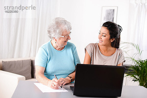 Fröhliche junge Frau  die einer älteren Person bei der Nutzung eines Laptops für die Internetsuche und E-Mail hilft