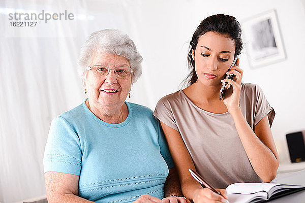 Fröhliche junge Frau  die einer älteren Person beim Papierkram und Telefonieren hilft