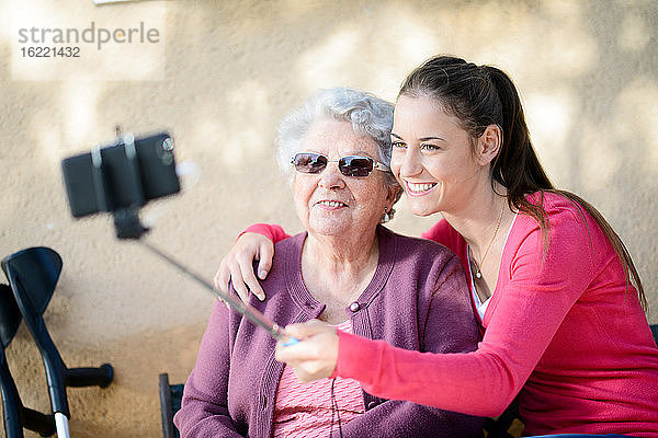 Fröhliche junge Frau macht ein Selfie mit einer älteren Frau in einem Garten eines Seniorenheims.