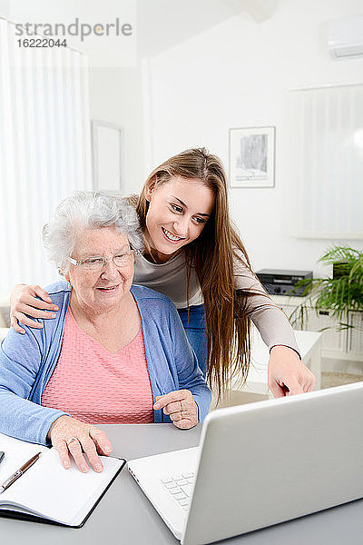 Junge Frau  die einer älteren Frau bei der Erledigung von Papierkram und Verwaltungsverfahren mit einem Laptop zu Hause hilft