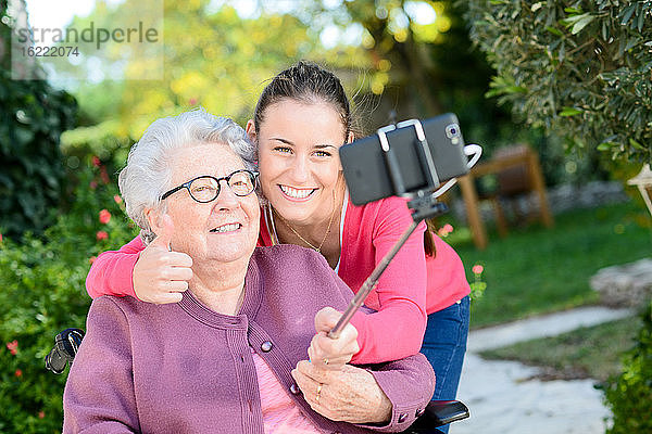 Fröhliche junge Frau macht ein Selfie mit einer älteren Frau in einem Garten eines Seniorenheims.