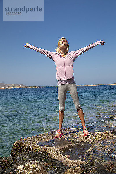 Eine schöne junge Frau in Sportkleidung  die sich in der Sonne vor dem Meer dehnt und die Luft einatmet.