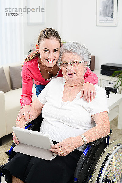 Fröhliches junges Mädchen  das mit einem Tablet-Computer im Internet spielt und Zeit mit einer alten Seniorin im Rollstuhl verbringt