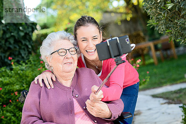 Fröhliche junge Frau macht ein Selfie mit einer älteren Frau in einem Garten eines Seniorenheims.