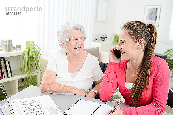 Junge Frau hilft einer älteren Frau bei der Erledigung von Papierkram und Verwaltungsaufgaben mit einem Laptop zu Hause