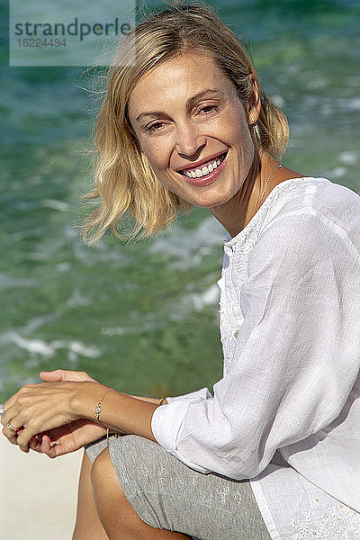 Schöne lächelnde junge Frau  die vor dem Meer sitzt.