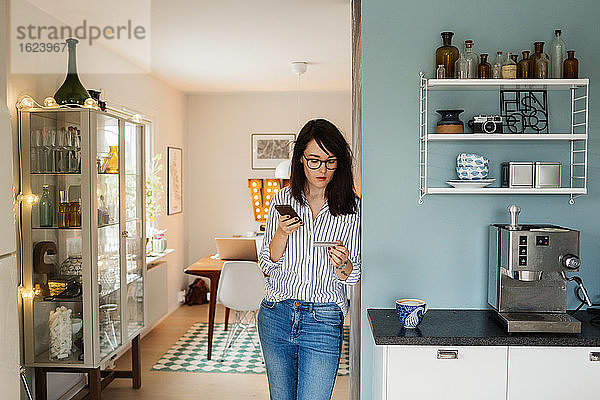 Junge Frau mit Smartphone und Kreditkarte in der Küche