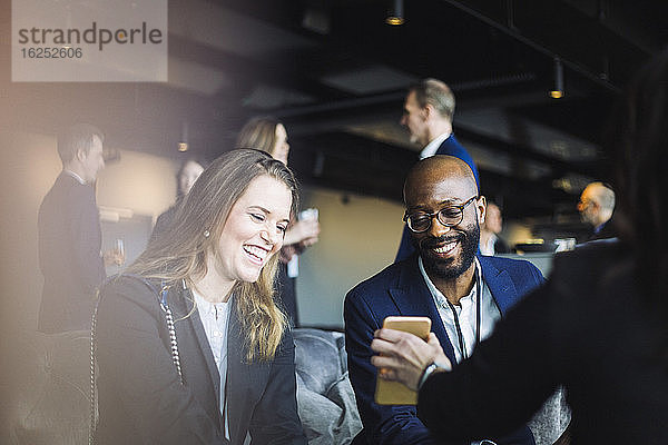 Lächelnde männliche und weibliche Mitarbeiter schauen auf das Mobiltelefon  während sie mit dem Unternehmer im Büro sitzen
