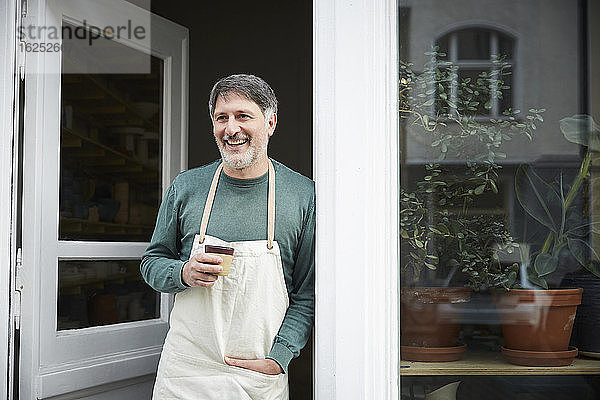 Lächelnder reifer männlicher Besitzer schaut weg  während er beim Kaffee an der Tür des Kunstateliers einen Kaffee trinkt