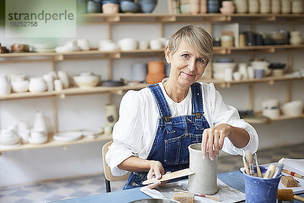 Porträt einer lächelnden reifen Frau mit Steingut bei Tisch im Kunstatelier