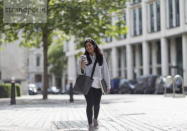 Lächelnde Frau mit Kopfhörern und Smartphone auf sonnigem Bürgersteig