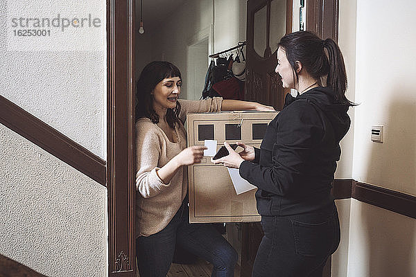 Lächelnde Zustellerin zeigt dem Kunden das Telefon  während sie eine Visitenkarte an der Türschwelle hält