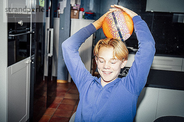 Lächelnder Junge hält Sportball in der Küche auf dem Kopf