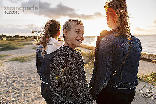 Lächelnder Teenager schaut weg  während er mit Mutter und Tochter am Strand steht