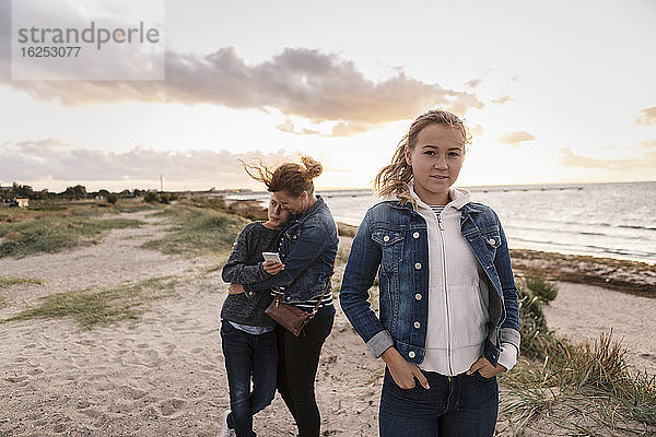Porträt eines Teenager-Mädchens  während die Mutter den Sohn am Strand umarmt