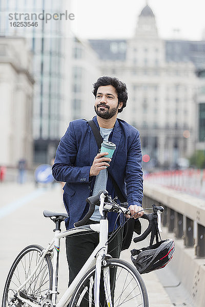 Lächelnder Geschäftsmann mit Kaffee und Fahrrad auf der Stadtbrücke