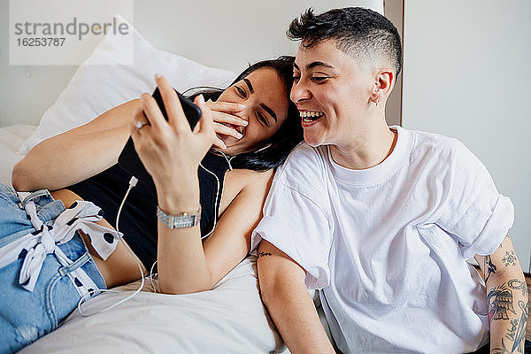 Junges lesbisches Paar in einem Schlafzimmer  schaut auf ein Handy  lächelt.