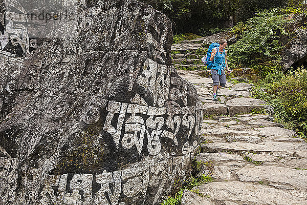 Eine Frau mit Rucksack geht einen Steinpfad entlang  streicht über eine Felswand mit Schriftzug; Nepal