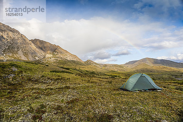 Ein Regenbogenbogen über einem grünen Rucksackzelt  das in der Tundra aufgestellt ist  Berge im Hintergrund an einem sonnigen Sommertag in der Arktis  Brooks Range  Arctic National Wildlife Refuge; Alaska  Vereinigte Staaten von Amerika
